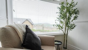 En beige lænestol med sort pude, et rundt bord og en potteplante ved et vindue med persienner.