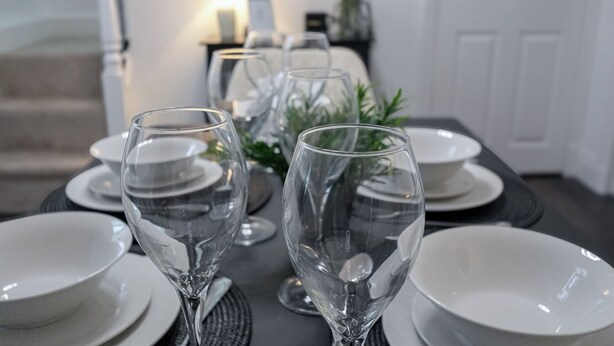 Et spisebord dækket med klare glas og hvide tallerkener, med en trappe og en dør i baggrunden.