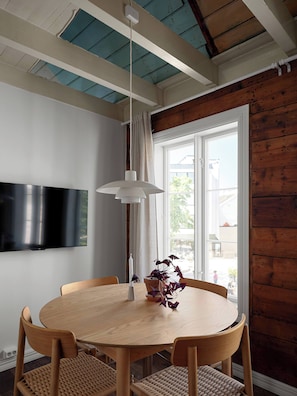 Een eethoek met een ronde tafel, vier stoelen, een hanglamp, een flatscreen televisie aan de muur en een raam met gordijnen.