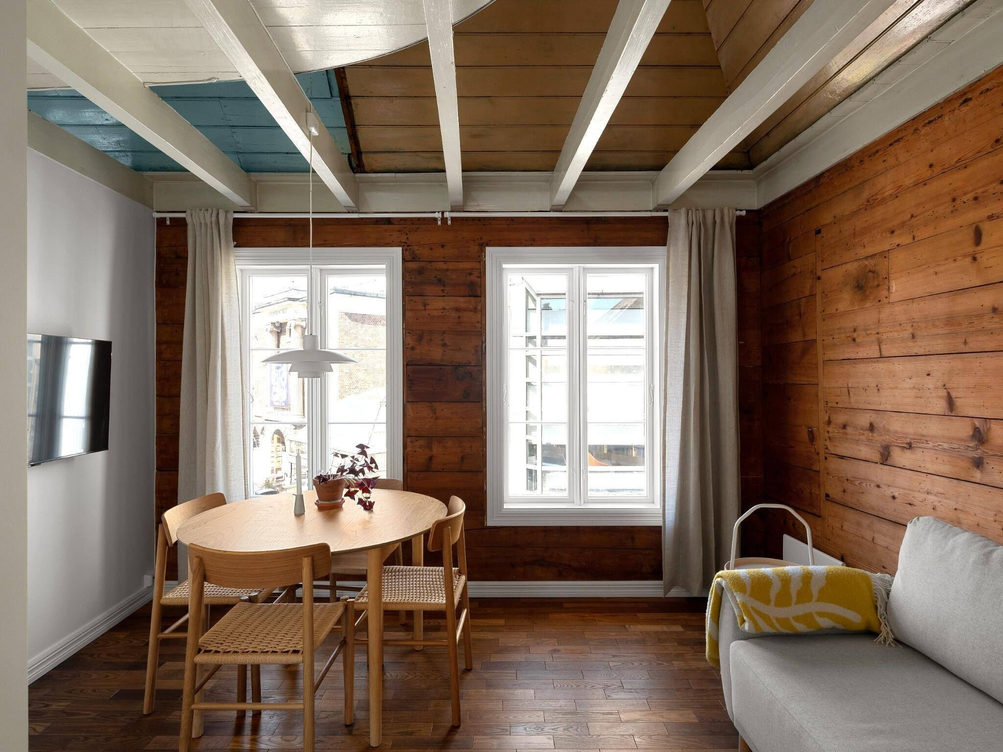 A room with a wooden ceiling, a round table with chairs, a sofa, and large windows.
