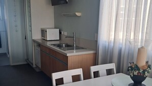A compact kitchen with a microwave, sink, and cabinets, a dining area with a table and chairs, and a window with sheer curtains.