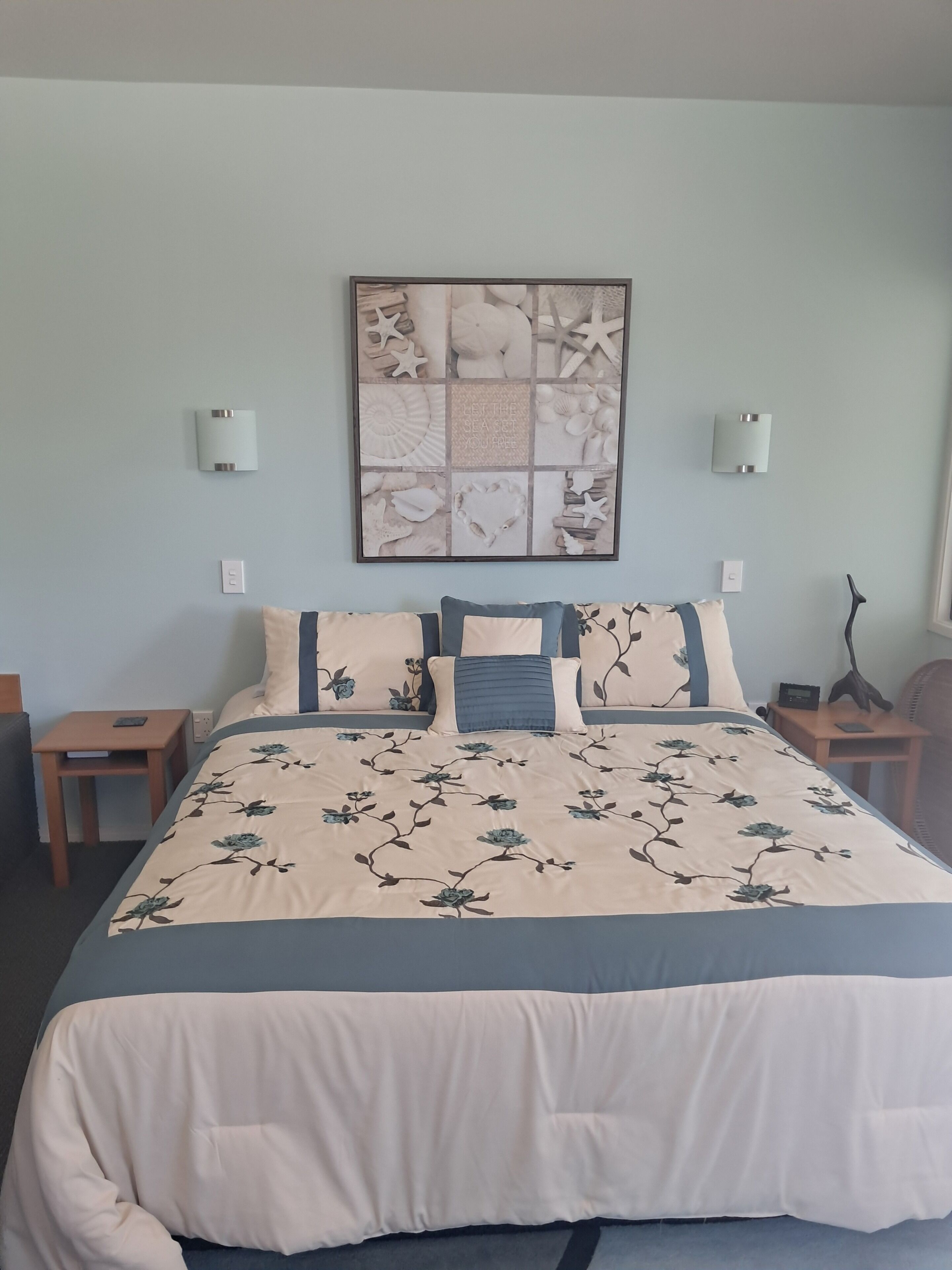 Een keurig opgemaakt slaapkamer met een bed, nachtkastjes, een kunstwerk aan de muur en een raam.
