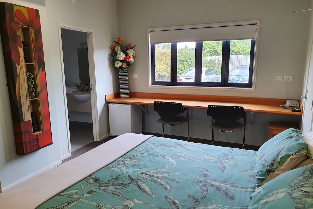 Un dormitorio con cama, escritorio, sillas, una ventana y un baño visible al fondo.