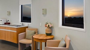 A modern room with a kitchen area, a dining table, two chairs, a vase with flowers, and a large window with a sunset view.