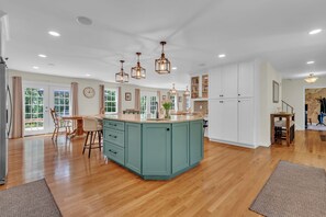 A spacious kitchen with a central island, pendant lighting, and a view of the outdoors through large windows. - The Harness Creek House (Annapolis)