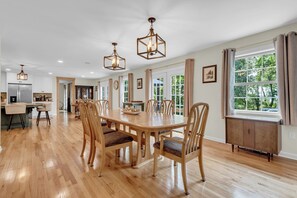 A spacious dining area with a wooden table and chairs, a kitchen in the background, and large windows with curtains. - The Harness Creek House (Annapolis)