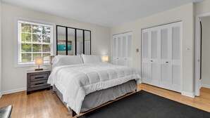 Quarto com cama, mesa de cabeceira, janela com persianas, espelho e um conjunto de armários brancos.
