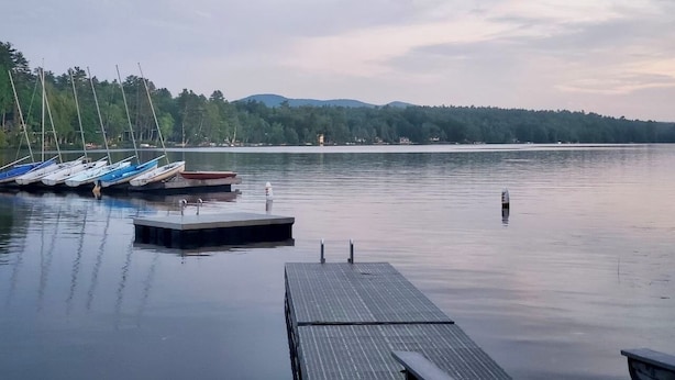 A calm lake with boats docked at a pier, surrounded by trees and hills.