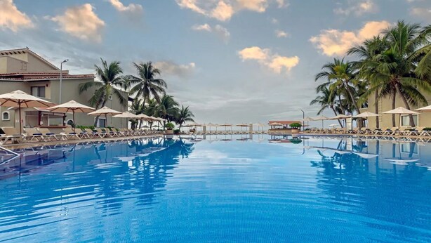 A swimming pool with lounge chairs and umbrellas, surrounded by palm trees and a building with a tiled roof. - Condominios Torre Continental Ixtapa (Ixtapa)