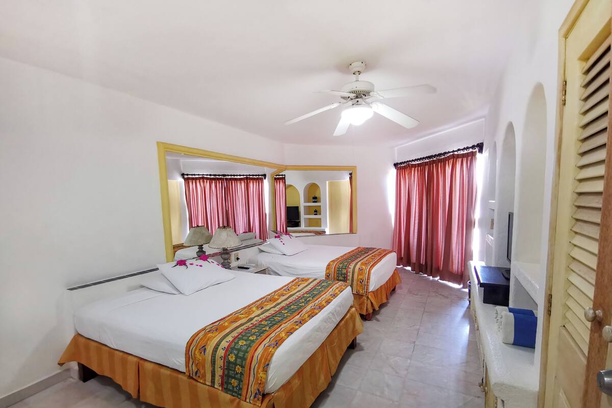 Habitación de hotel con dos camas, ventilador de techo, espejo, televisor y puerta de baño.