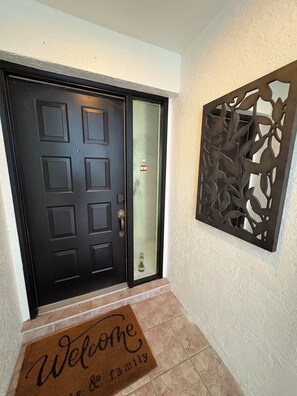 Une porte noire avec un panneau de verre, un paillasson et un panneau métallique décoratif sur le mur.