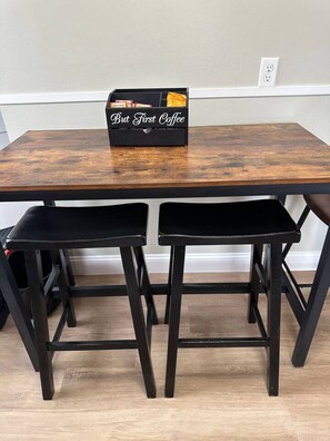 A wooden table with two black stools, a black box labeled 'But First Coffee,' and a white electrical outlet on the wall. - Close to U in Champaign, 2 Bedroom House (Champaign)