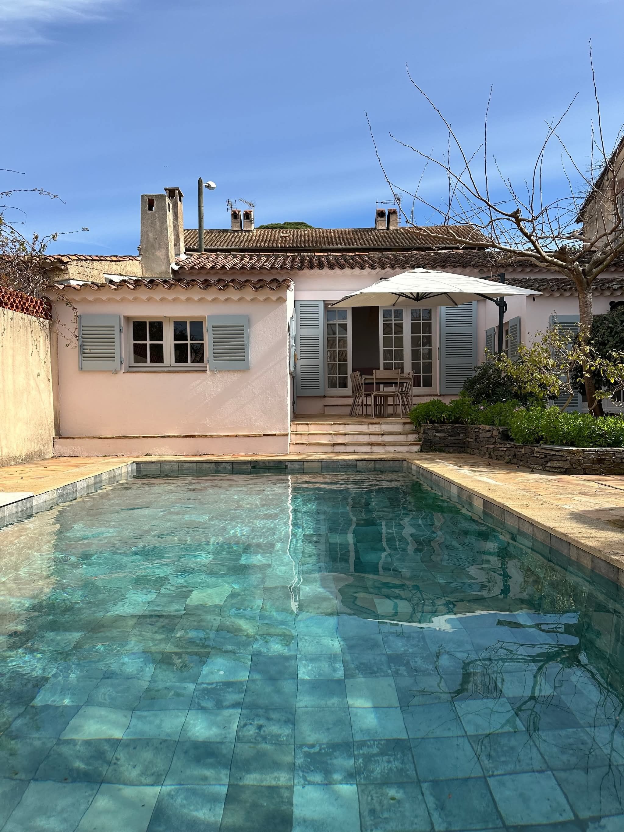 A swimming pool with clear water, a patio with outdoor furniture, and a building with a tiled roof and shutters.