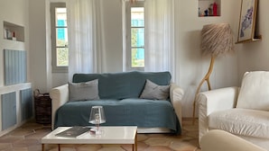Ett vardagsrum med en soffa, ett soffbord, en hängande lampa och utsikt över omgivningen genom ett fönster.