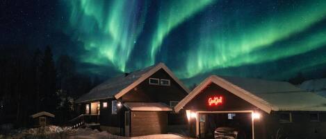 Una cabaña con garaje y un letrero de neón, frente a un cielo nocturno con auroras boreales.