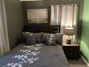 A bedroom with a bed, a nightstand, a lamp, a painting on the wall, and a window with curtains.