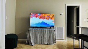 A living room with a flat-screen TV on a stand, a striped table, a black ottoman, and a door leading to another room. - Hidden Gem Sarasota Getaway Near Siesta Key (Sarasota)