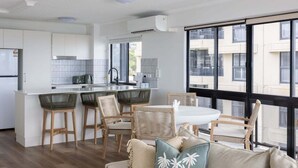 A modern kitchen with white cabinets, a dining area with wicker chairs, and a large window with a view of buildings. - Summer Cove - Beachfront with Pool (Sunshine Coast)