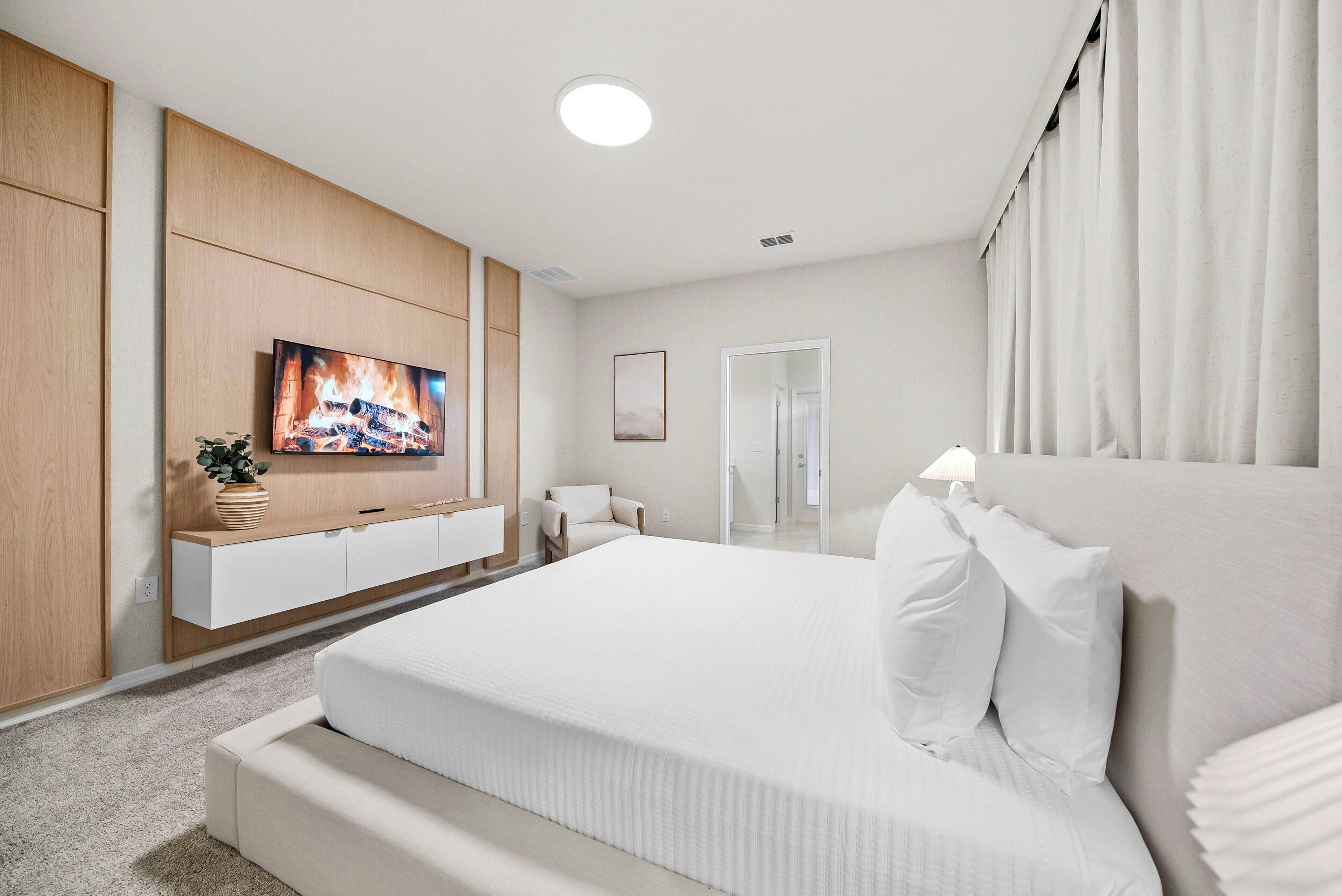 Quarto de hotel com uma cama grande, uma televisão presa a um painel de madeira, uma mesa de cabeceira com um vaso e uma porta que dá para um banheiro.