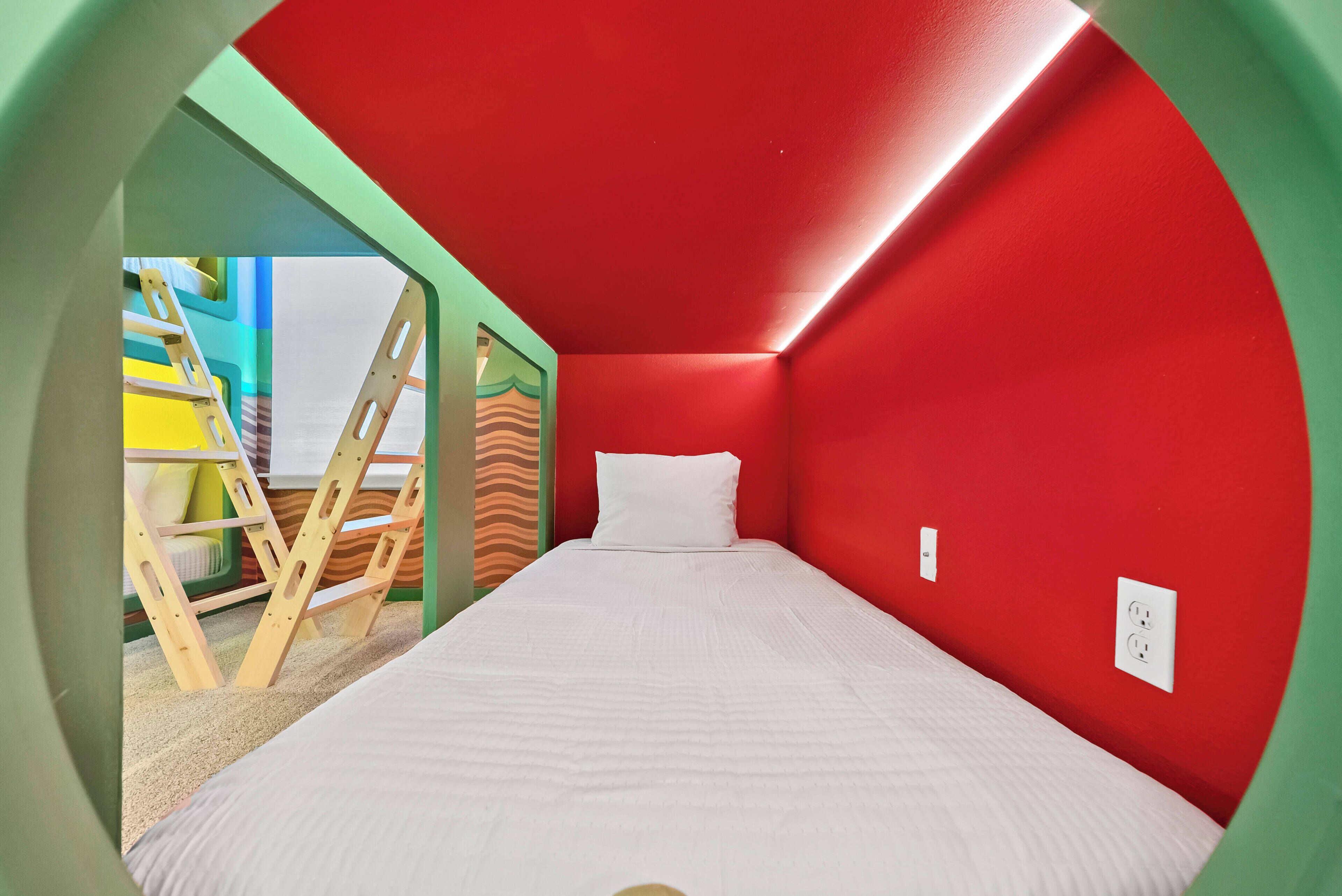 A room with a bed, a red wall, and a view of a colorful play area.
