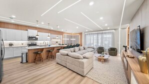 A modern kitchen with white cabinets, a microwave, and a coffee maker. There is a dining area with a large beige sofa and a coffee table. The room has wooden flooring and a large window with curtains. - 10BR house at Windsor Cay Resort (Clermont)