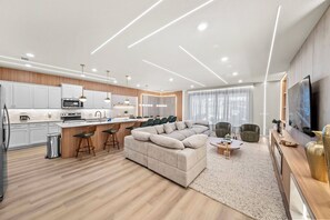 Eine moderne Küche mit weißen Schränken, einer Mikrowelle und einer Kaffeemaschine. Es gibt einen Essbereich mit einem großen beigen Sofa und einem Couchtisch. Der Raum verfügt über Holzböden und ein großes Fenster mit Vorhängen.
