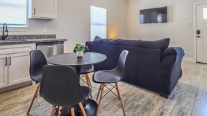 A modern living room with a dark sofa, a dining table with chairs, a ceiling fan, and a TV mounted on the wall.