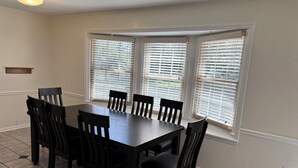 Un comedor con mesa y sillas de madera oscura, ventanas amplias con persianas y pared de color claro.