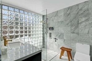 A bathroom with a glass shower enclosure, a marble-tiled wall, a wooden stool, and a white toilet. - 3BR oasis: private pool & mountain views (Palm Springs)