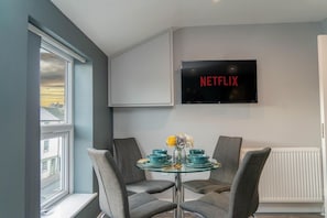 Ein modernes Zimmer mit Glastisch, grauen Stühlen, einem Wandfernseher und einem Fenster mit Aussicht.