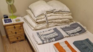 A single bed with pillows, folded blankets, and a folded towel. There is a bedside table with a lamp and a book.