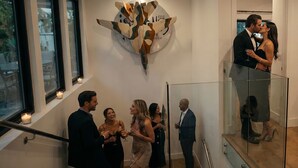 Un interior moderno con una gran obra de arte abstracta en la pared, una escalera con barandillas de vidrio y un grupo de personas reunidas.
