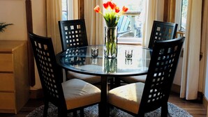 Un coin repas comprenant une table ronde en verre, quatre chaises, un vase avec des fleurs et une vue sur l’extérieur à travers une fenêtre ornée de rideaux.