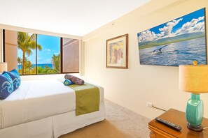 Ein Hotelzimmer mit Bett, Fenster mit Blick auf Palmen und Meer, ein Meeresmotiv als Bild und eine Lampe.