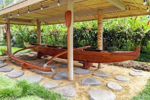 Ein überdachter Außenbereich mit zwei traditionellen Booten, einem Reetdach und einem Garten mit tropischen Pflanzen.