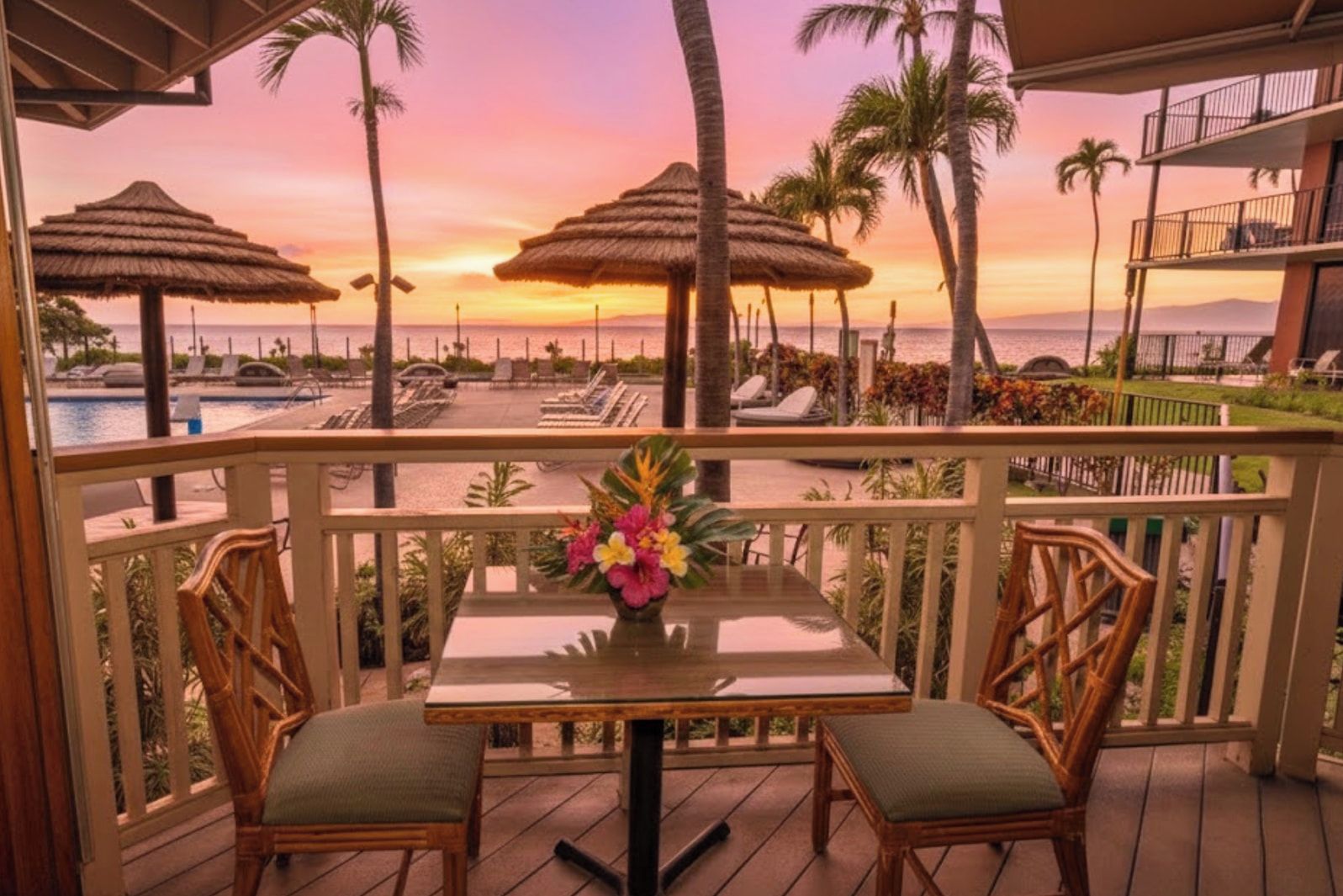 Un balcon avec une table et des chaises, donnant sur une piscine et une plage, agrémentée de parasols et de palmiers.