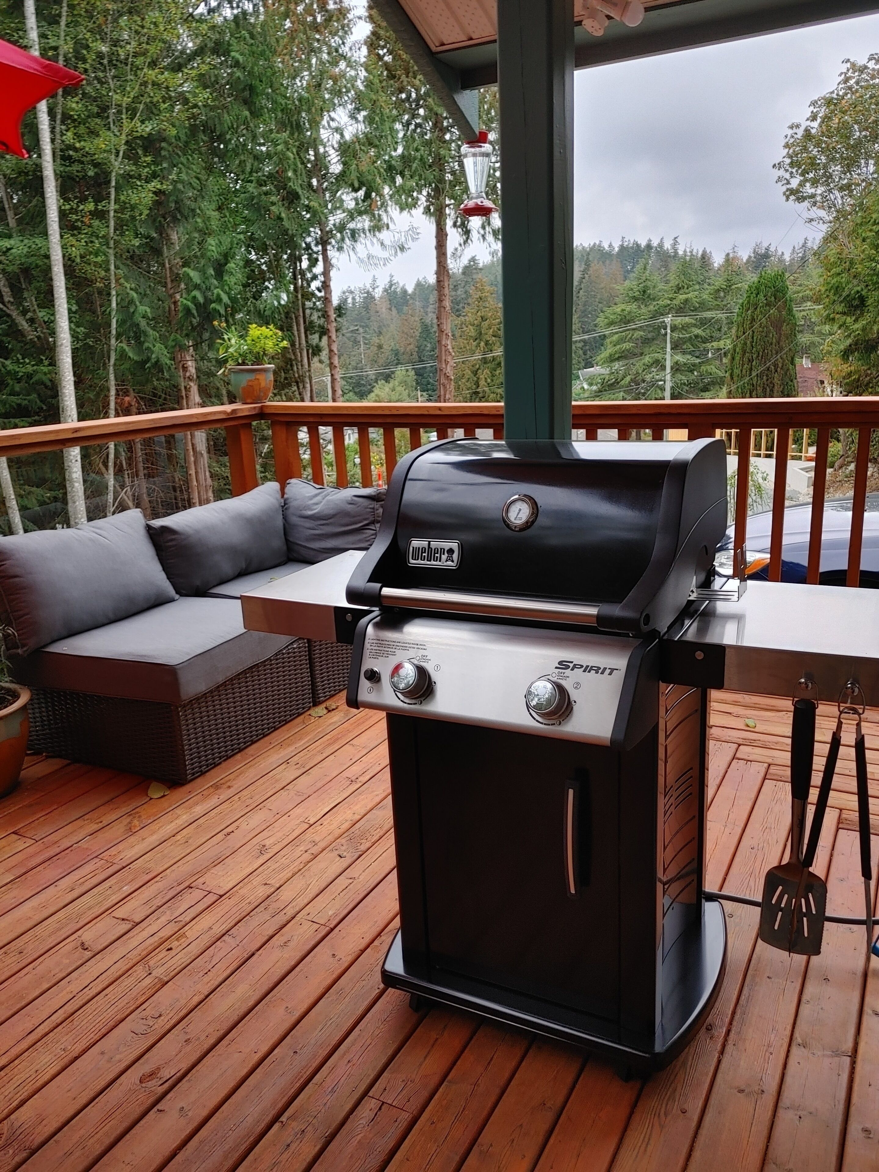 Ein Weber Spirit Grill steht auf einer Holzterrasse, mit einem grauen Sofa und einem roten Sonnenschirm.