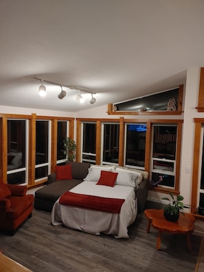 Un dormitorio acogedor con cama, sofá y una mesita. Amplias ventanas con marcos de madera, una lámpara de techo y una estantería con adornos.