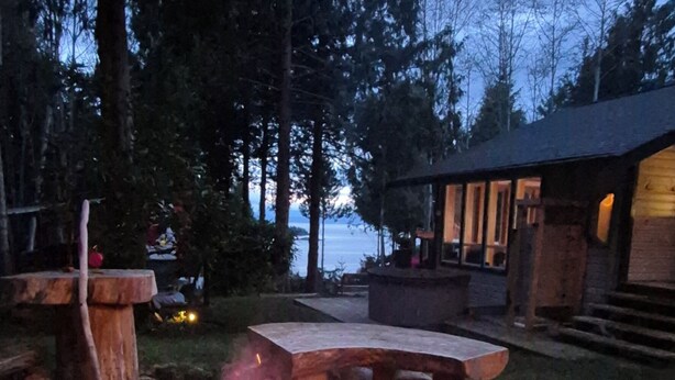 A circular fire pit with flames, surrounded by a brick structure, set in a wooded area with a building in the background. - SeaView Getaway- B.L.T. (bed and local treats), Retreat. (Sechelt)