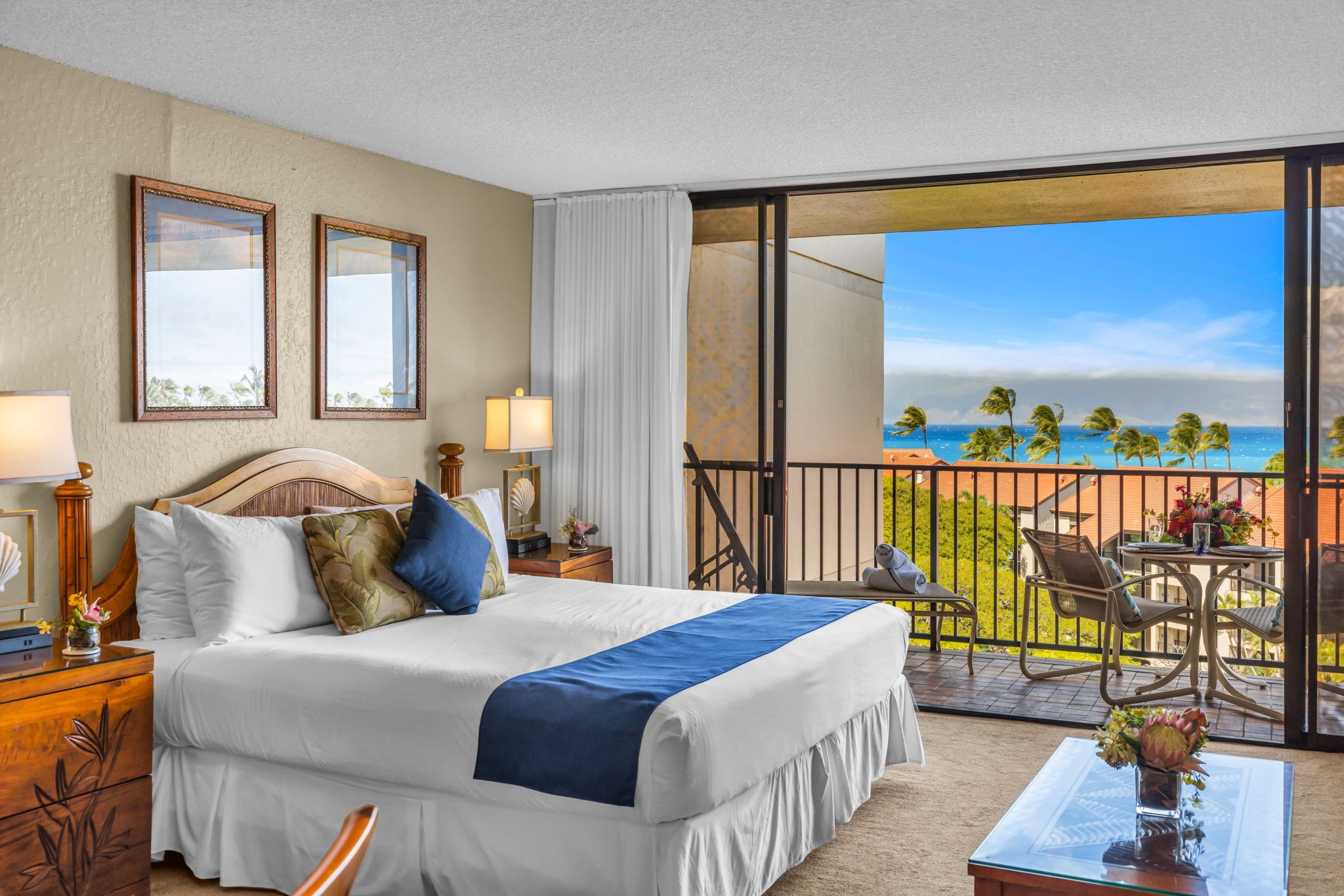 Habitación de hotel con cama, mesitas de noche, un balcón y vistas al océano y palmeras.
