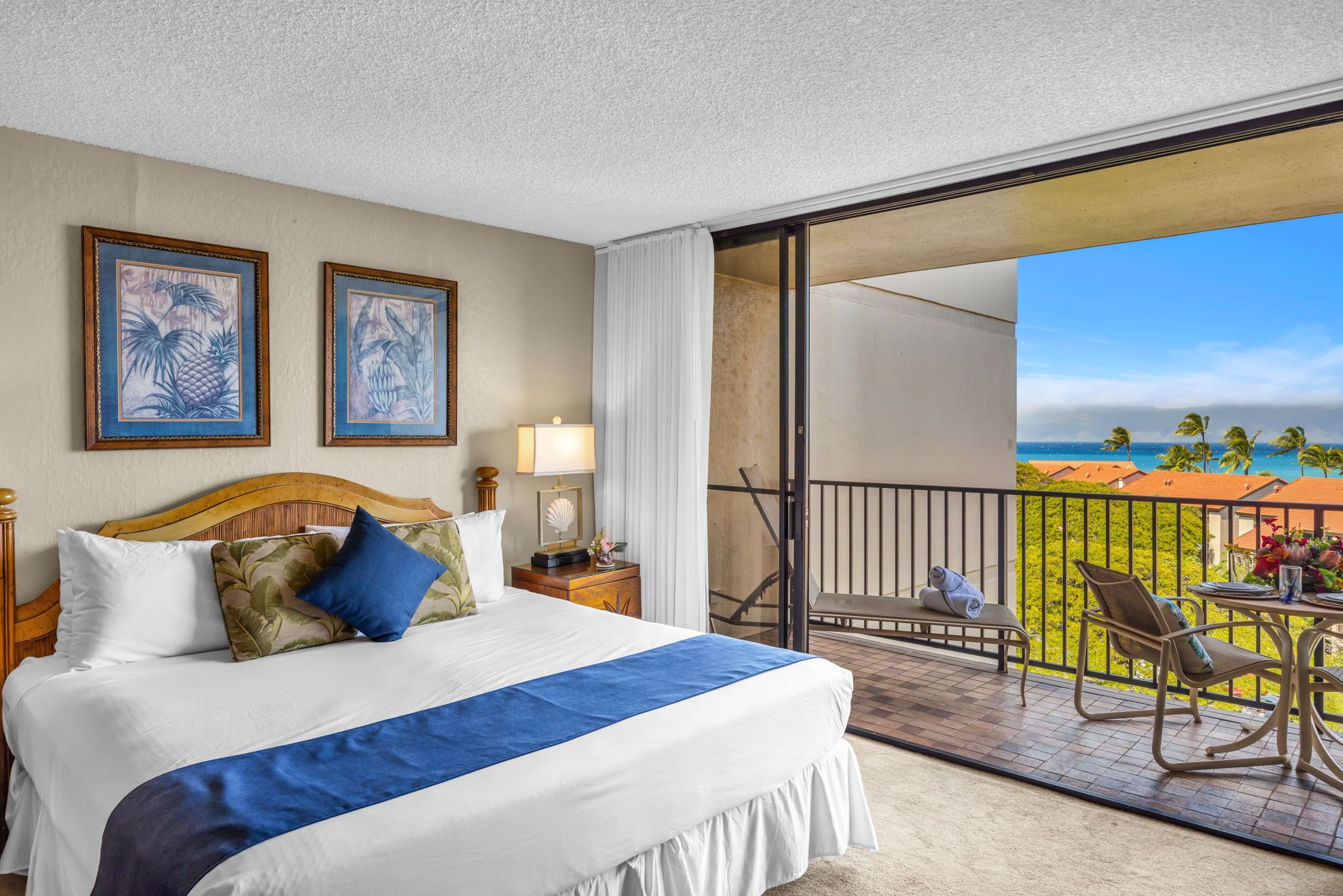 A hotel room with a bed, a nightstand, a lamp, a view of the ocean, and a balcony with chairs and a table.