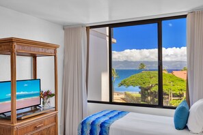 Ein Schlafzimmer mit einem großen Fenster, einem Fernseher auf einem Holztisch und einem Bett mit einer blau-weißen Tagesdecke.