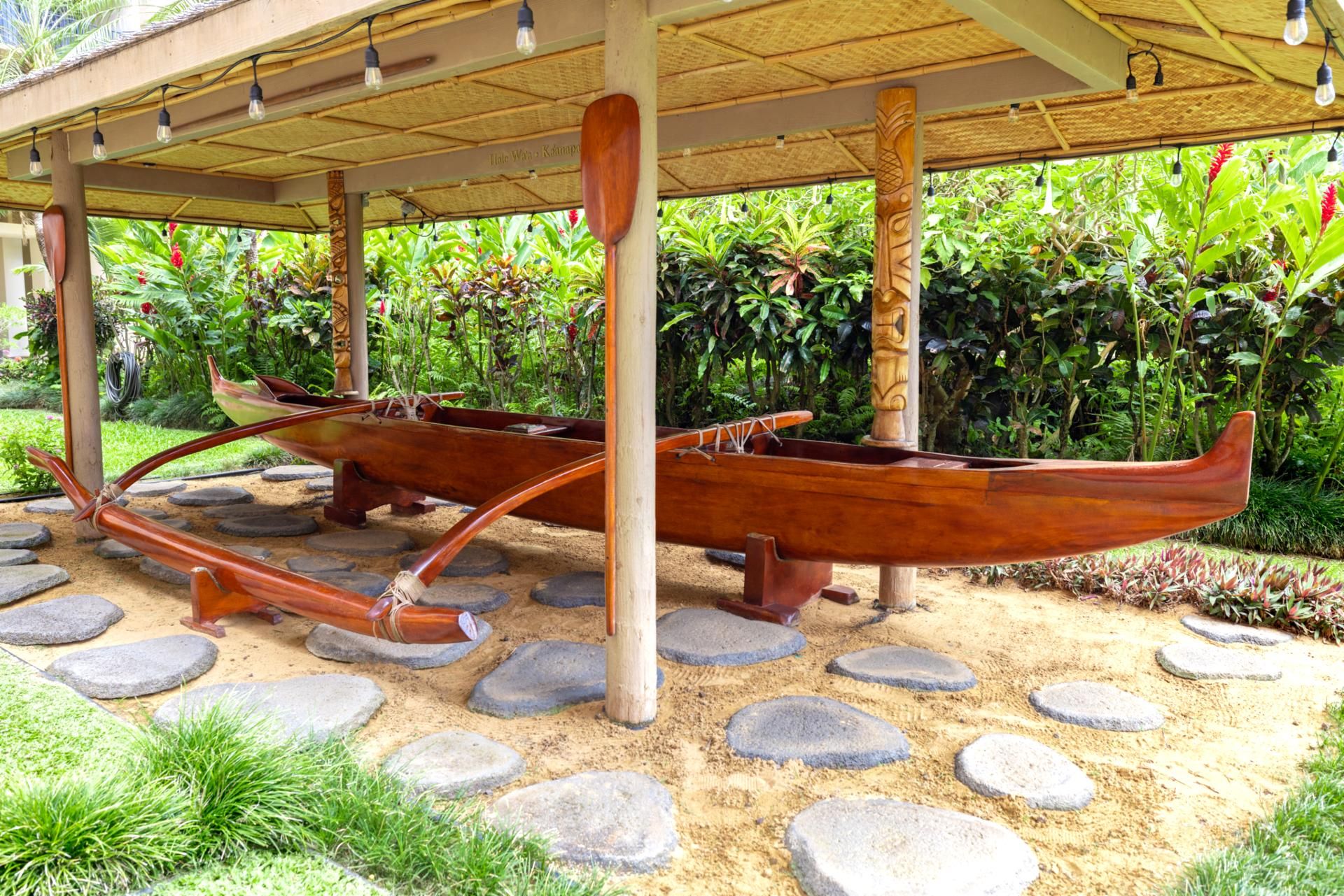 A traditional wooden boat under a thatched roof structure, surrounded by a garden with tropical plants and stone stepping stones.