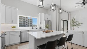 A modern kitchen with white cabinetry, a central island, black bar stools, and pendant lighting. - Capri-Lux Pool/Hot-Tub/Steps To the Beach (Treasure Island)