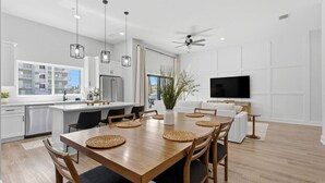 A modern kitchen with white cabinets, a dining area with a wooden table and chairs, a ceiling fan, and a TV mounted on the wall. - Capri-Lux Pool/Hot-Tub/Steps To the Beach (Treasure Island)