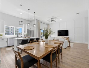 Eine moderne Küche mit weißen Schränken, eine Essecke mit Holztisch und -stühlen, ein Deckenventilator und ein an der Wand befestigter Fernseher.