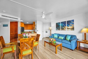 A living room with a blue sofa, wooden chairs, a coffee table with flowers, a ceiling fan, and a kitchen in the background. - Kaanapali Shores | Ocean View 7 BR, Sleeps 22 | Car Incl. w/ 6+ Nights | KS ML-3773 by KBM (Lahaina)