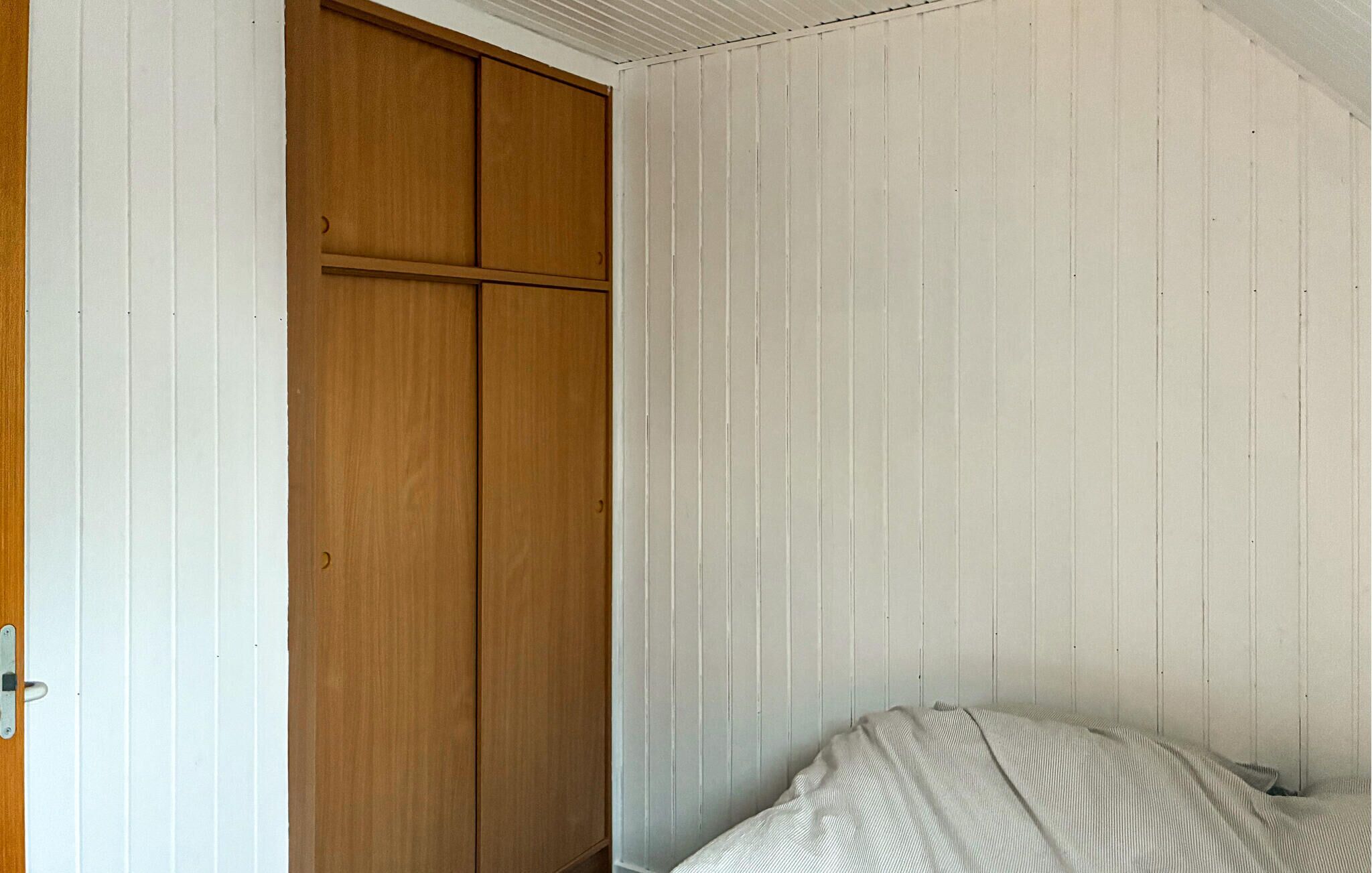 Ein Schlafzimmer mit einem Holzschrank, einem Bett mit einer Streifenoptik und weiß geteilten Wänden.