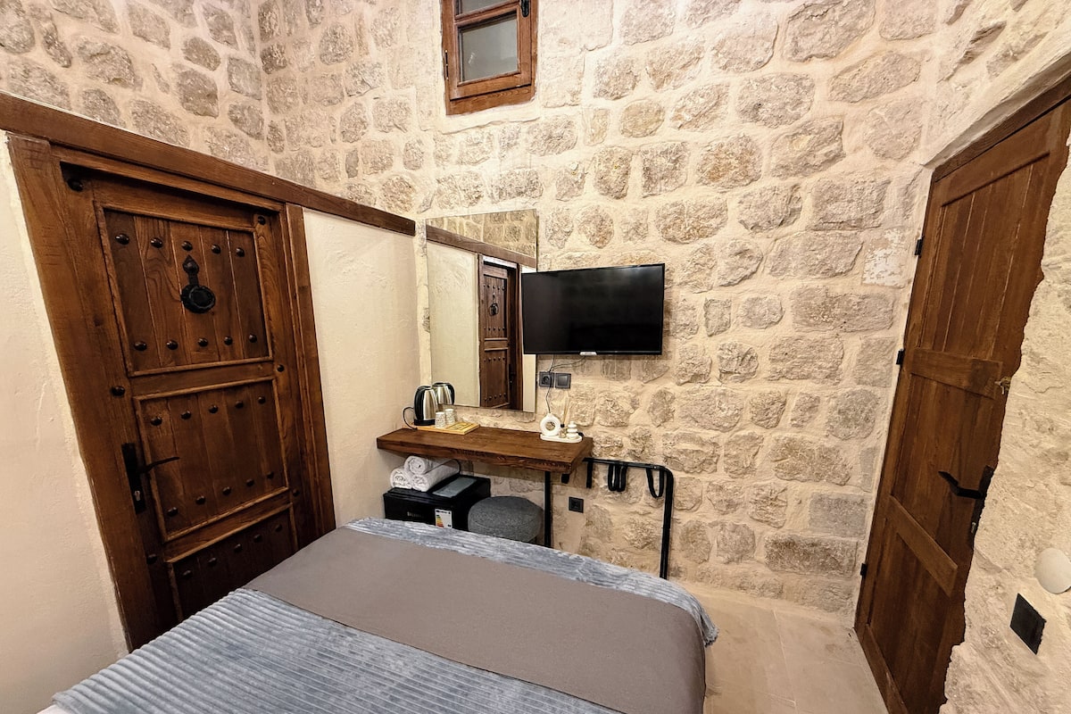 A room with a bed, a wooden wardrobe, a desk with a mirror, a television, and stone walls.