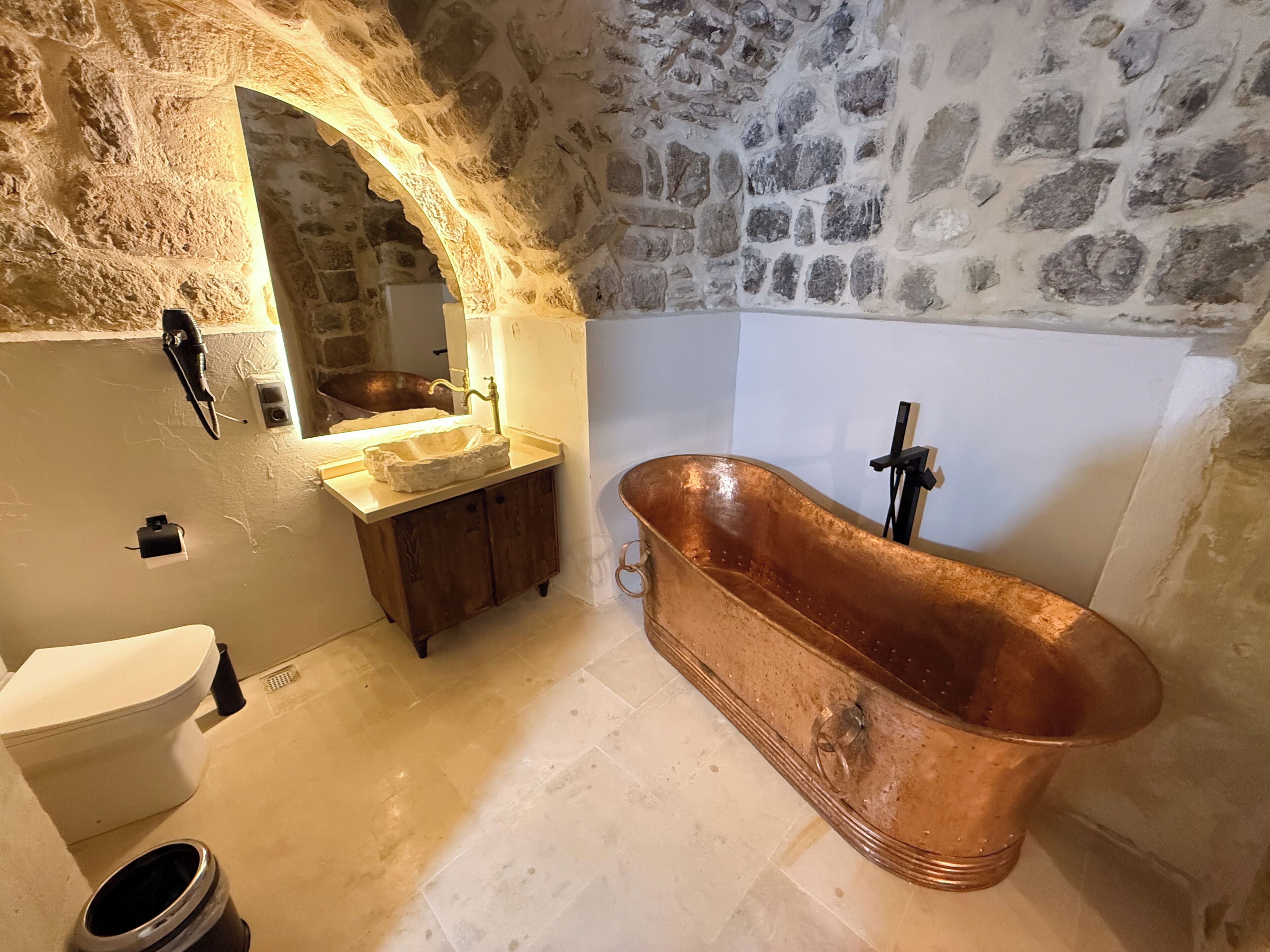 A bathroom with a stone wall, a copper bathtub, a sink with a mirror, and a toilet.